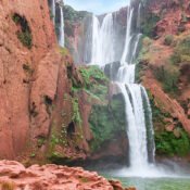 A powerful waterfall plunges down red cliffs into a calm, green pool below